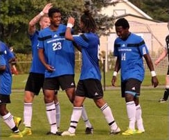 Four soccer players in blue jerseys celebrate on the field, with one player giving a high-five.
