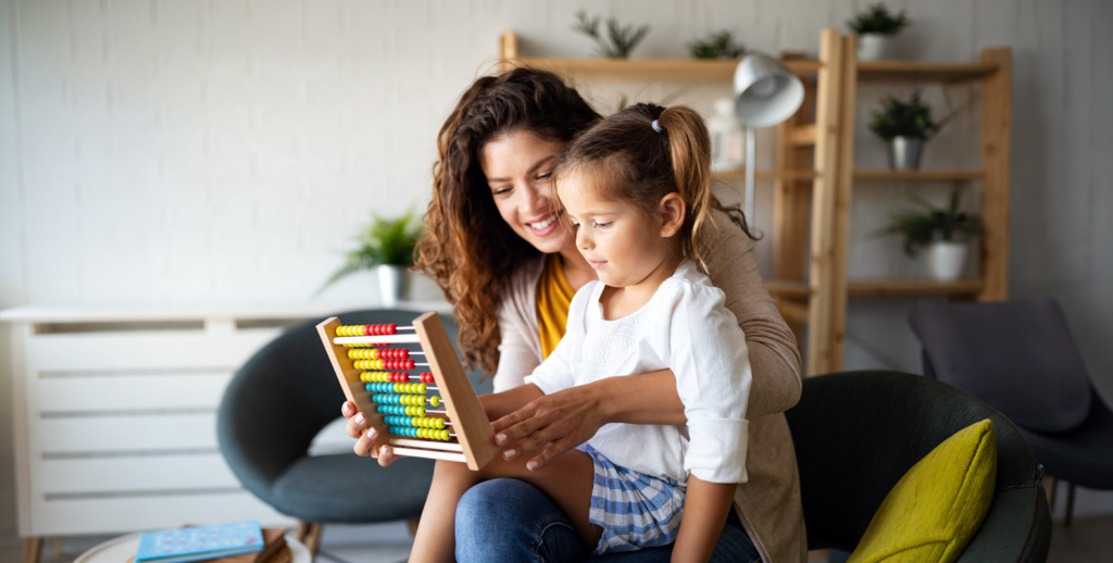 A woman and a young girl sit together on a chair, smiling and using a colorful abacus—a wonderful scene that reflects the joy of Early Childhood Education—in a bright living room with shelves and plants in the background.