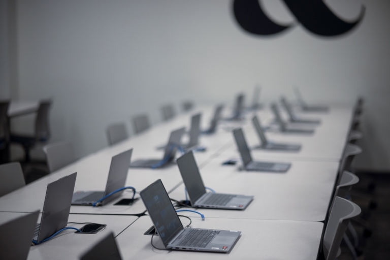 A row of open laptops on a long white table in an empty, modern classroom or office with gray chairs and a partial black wall design visible.