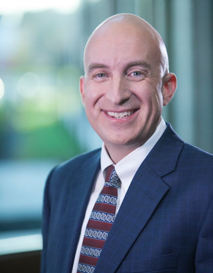 Smiling bald man in navy suit and patterned tie standing in front of blurred background.