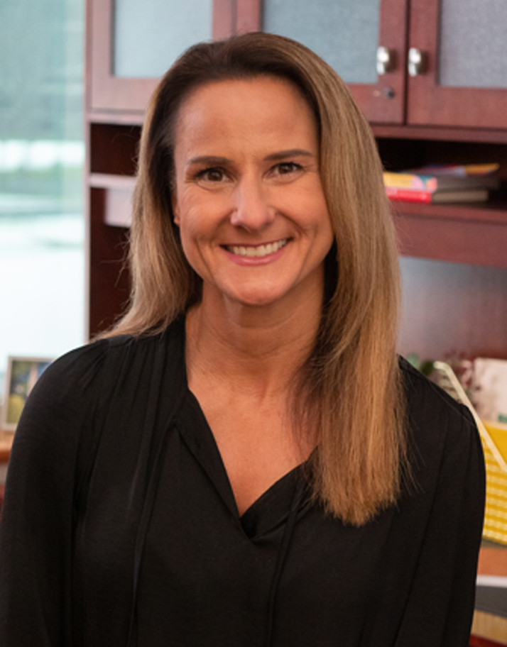 Dr. Laura Voelkl, with straight, shoulder-length blonde hair and a black top, smiles while sitting in an office setting with bookshelves and cabinets in the background.