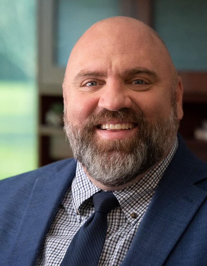 A bearded man in a blue suit and tie smiles at the camera with a blurred background.