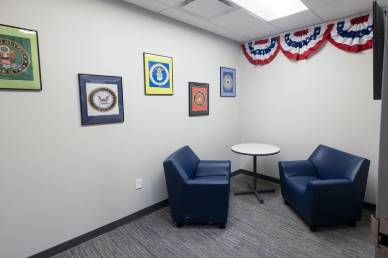 Small room with two blue armchairs facing a round table, military service emblems framed on the wall, and red, white, and blue bunting hanging above. Neutral walls and gray carpeted floor.