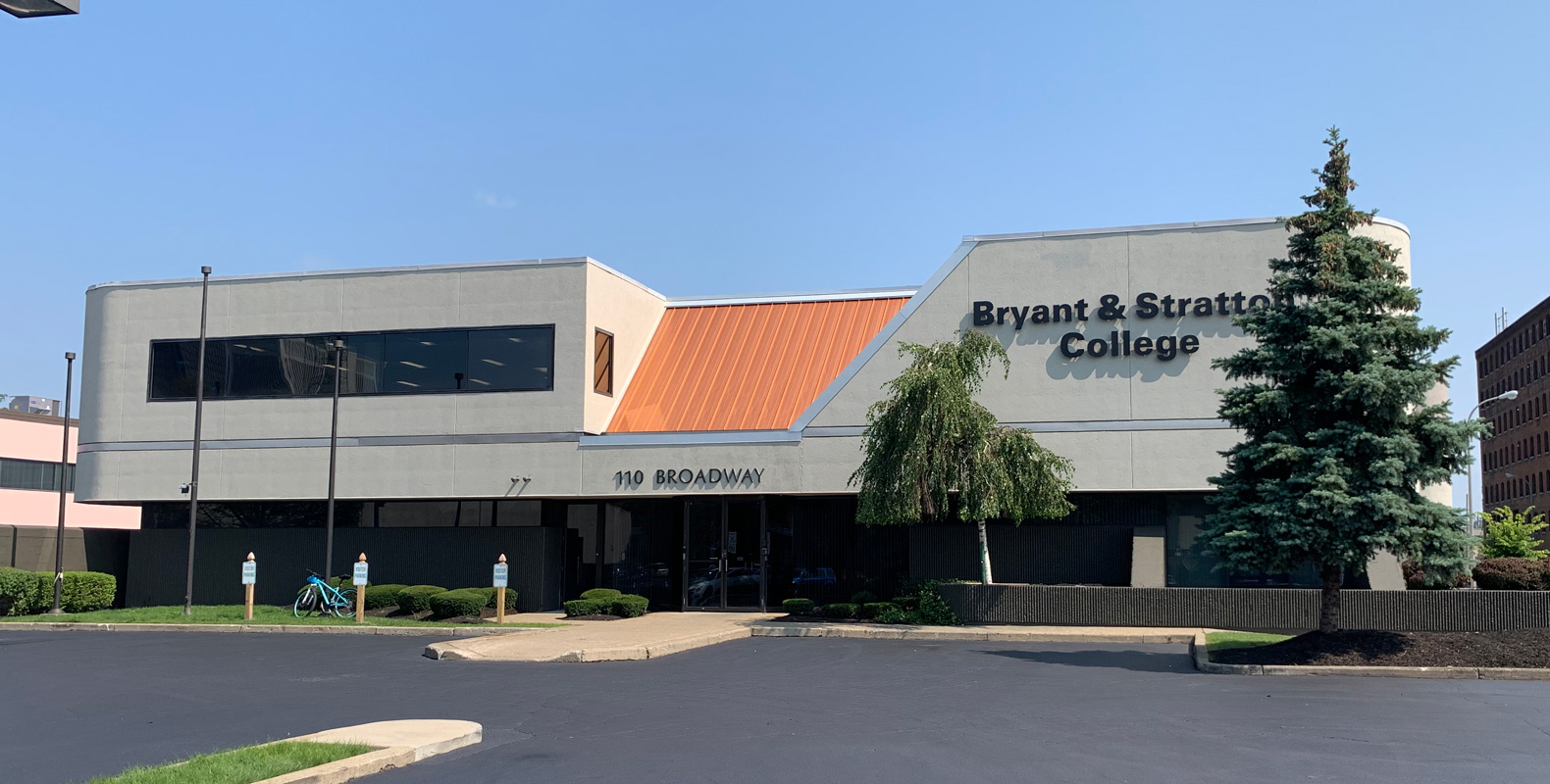 A modern two-story building with an orange roof section and large windows displays the sign "Bryant & Stratton College" on the front. The entrance is labeled "110 Broadway," with trees and a parking lot in front.