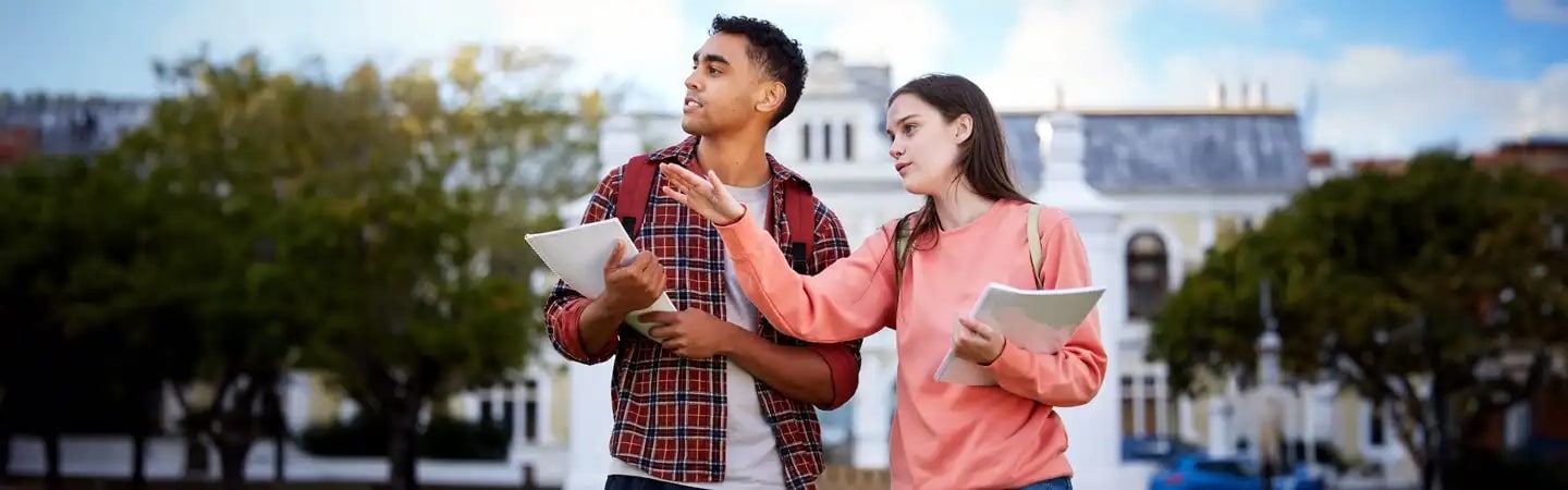 Two students with backpacks hold tablets and papers, discussing something outside in a park-like area with buildings behind them.