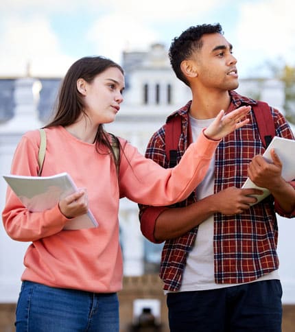 Two students stand outdoors, holding notebooks and backpacks. The female student, wearing a peach sweatshirt, gestures with her hand. The male student, in a plaid shirt, listens attentively. A building is visible in the background under a blue sky.