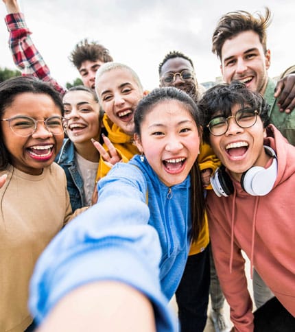 A group of seven diverse young people taking a cheerful selfie outdoors. They are smiling and wearing casual clothing, with the person in front holding the camera. The sky is slightly cloudy in the background.