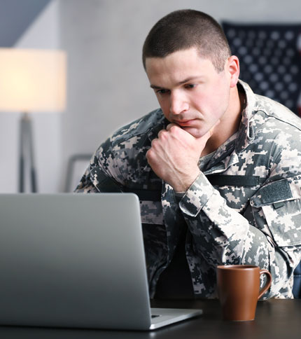 A person in a digital camouflage military uniform sits at a desk, looking intently at a laptop screen. A brown mug is on the desk in front of them. A lamp and a partially visible flag are in the background.