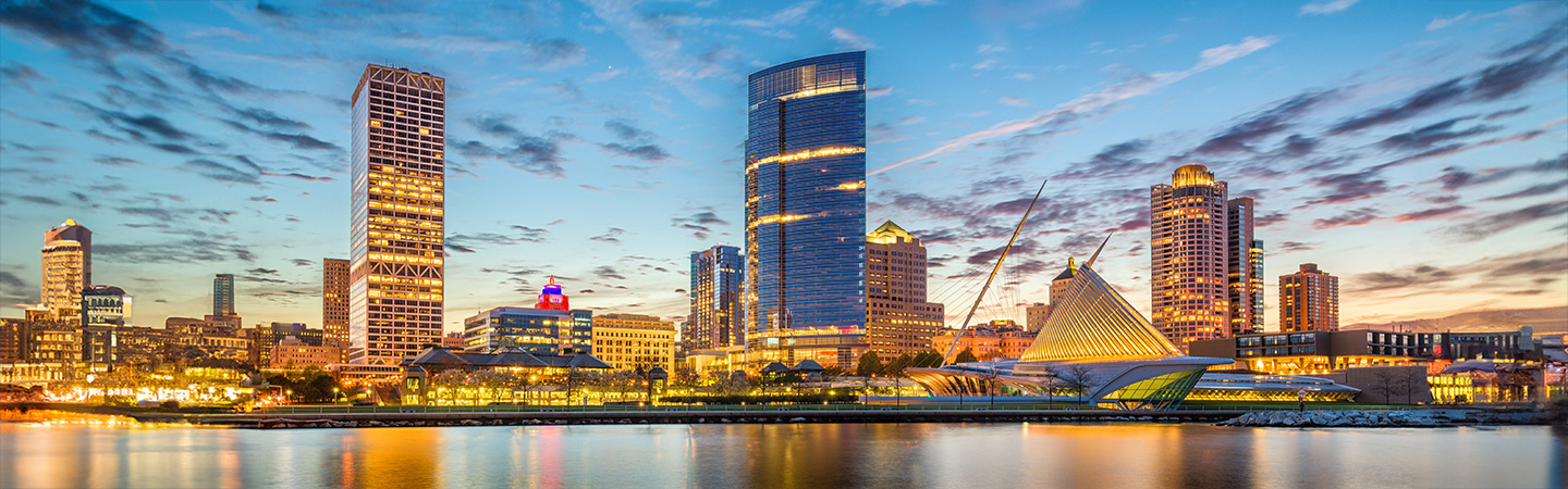 A panoramic view of a city skyline at dusk, featuring tall buildings reflecting on the water.