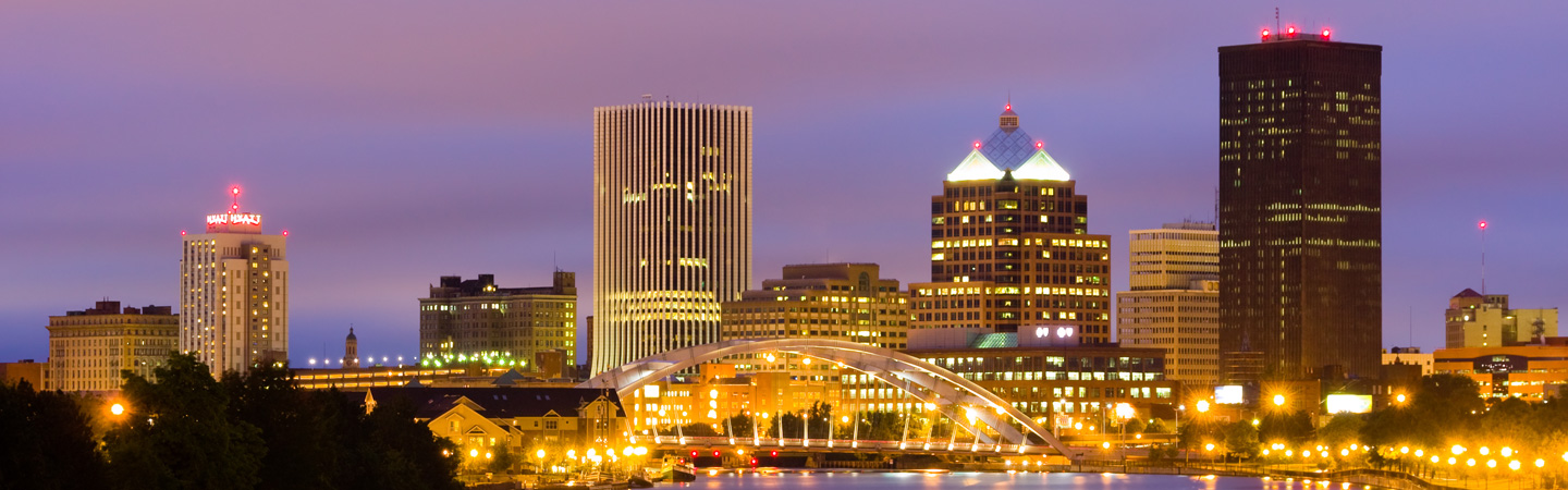 Nighttime cityscape of Rochester, NY, featuring bright city lights, a lit-up bridge, and tall buildings against a dusky sky.