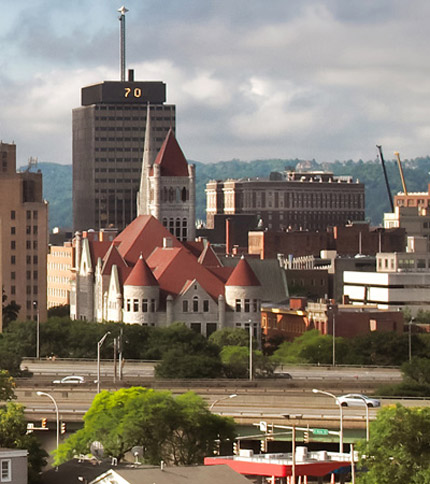 The Syracuse skyline with prominent buildings and lush greenery under a partly cloudy sky.