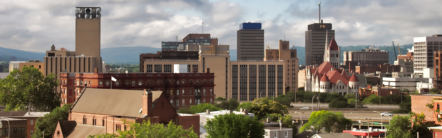 A city skyline featuring various buildings, a prominent clock tower, and a scenic backdrop of trees and hills.