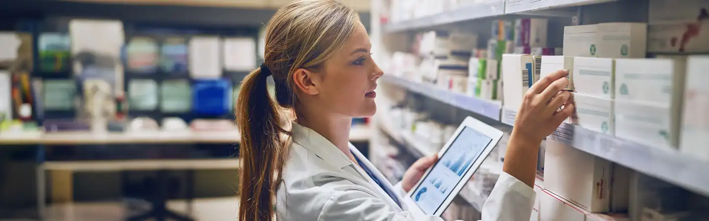 A woman in a lab coat holds a tablet and inspects medication boxes on a pharmacy shelf.