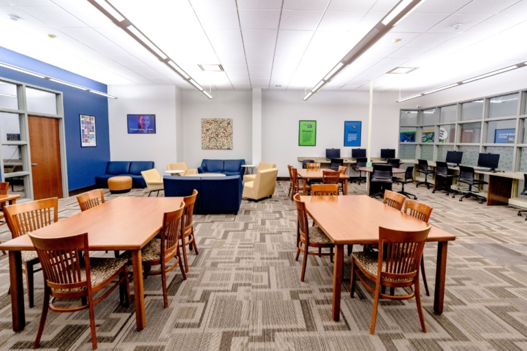 A modern library room with wooden tables and chairs, blue and beige sofas, computers along the wall, large windows, and colorful posters on blue and white walls. The space is well-lit with ceiling lights.
