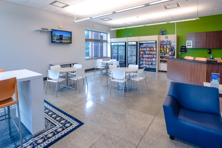 A bright, modern waiting area with tables, chairs, a blue couch, vending machines, a TV on the wall, and a counter with brochures. Large windows let in natural light. The floor is polished concrete.