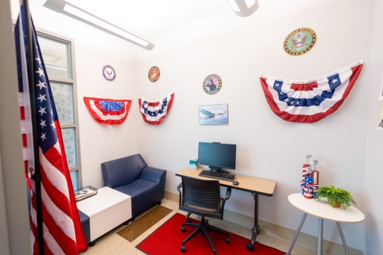 Small office decorated with patriotic bunting, military emblems on the wall, and an American flag. There’s a desk with a computer, a chair, a blue armchair, and a small table with patriotic decorations.