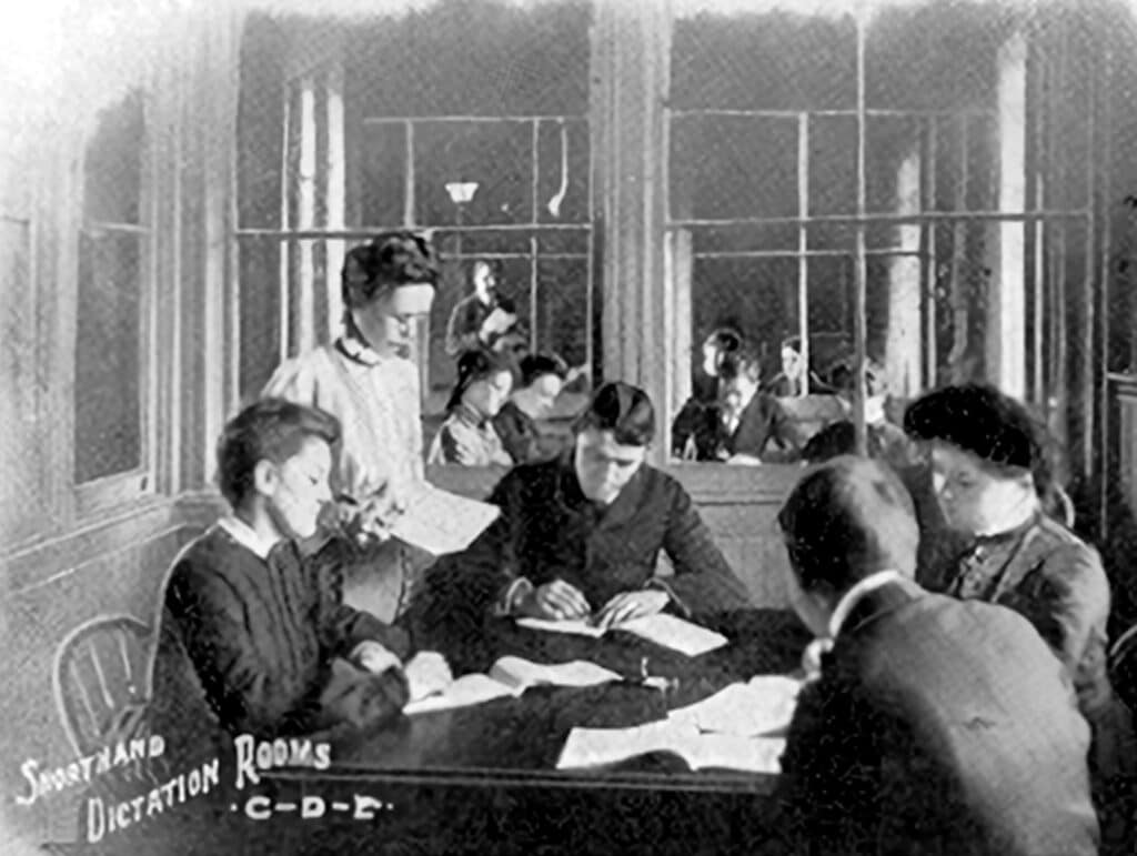 A vintage photo of a group of people in a classroom practicing shorthand and dictation with books and papers.