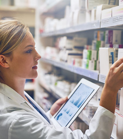 A pharmacist in a white coat is standing in front of shelves filled with medication boxes. She is holding a tablet and checking a box of medicine, appearing focused on her task. The background is lined with a variety of colorful medicine packages.