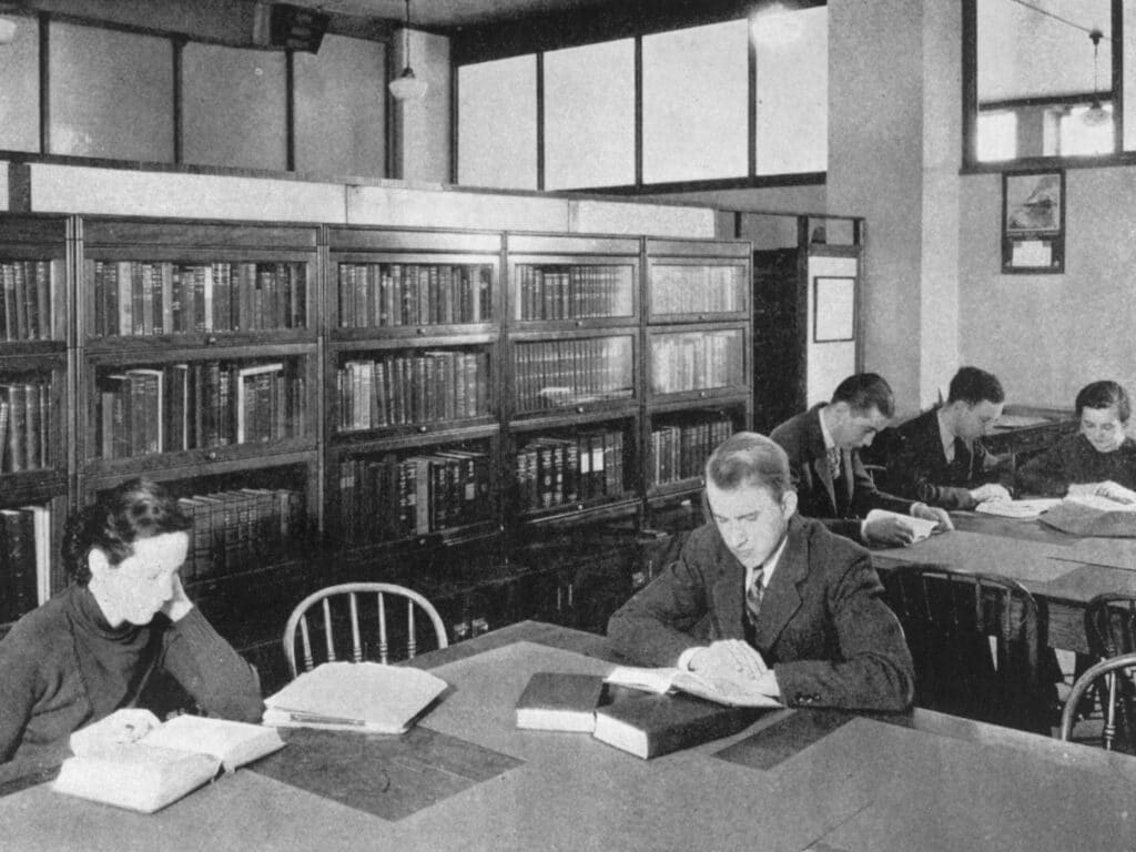 People read books at tables in a quiet library with rows of bookshelves in the background.