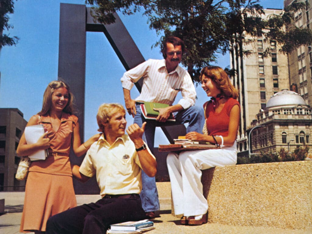 A group of four young adults study and chat outside near a large metal sculpture and buildings in the background.