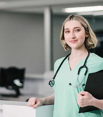 A person wearing light green scrubs and a stethoscope, likely having pursued an associate degree in nursing, stands confidently in a hospital or clinic setting, holding a black folder. The background features a reception desk and office equipment, slightly blurred.