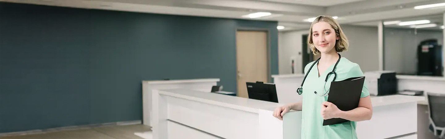 A nurse in scrubs stands with a clipboard in a modern, brightly lit hospital reception area.
