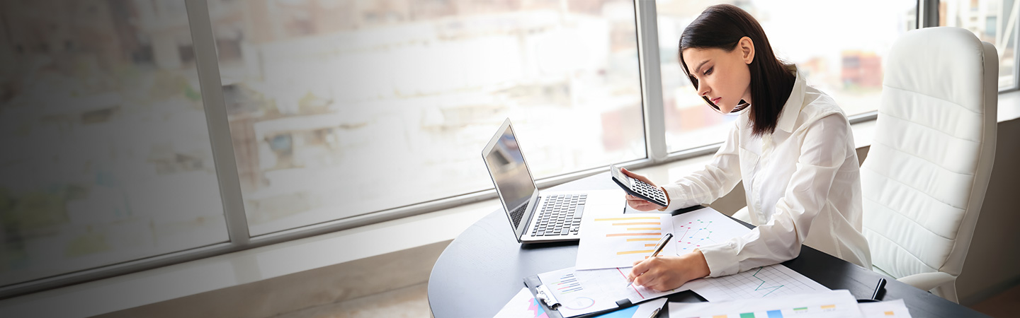 A woman multitasks in an office, holding a phone and calculator while writing, with a laptop and charts on the desk.