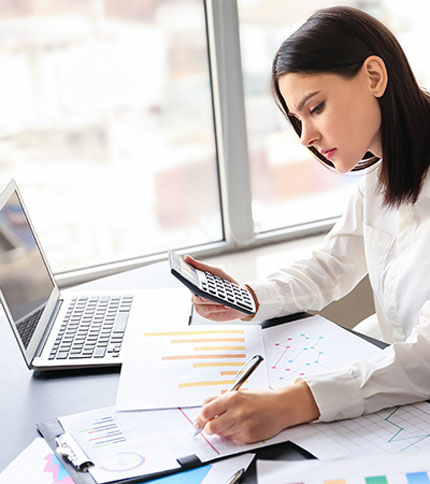 A woman sits at a desk, focused on calculating figures with a calculator. She has various graphs and charts in front of her and a laptop open on the desk. She wears a white blouse, and large windows are in the background.