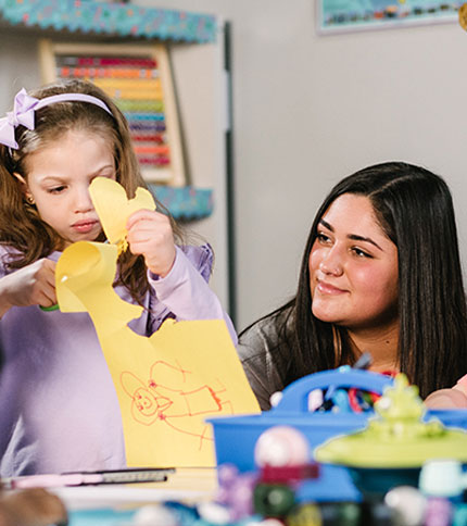 A young girl in a purple shirt uses scissors to cut a yellow paper with a drawing of a person on it. An adult woman sits beside her, smiling. They are in a classroom setting with colorful supplies around them.