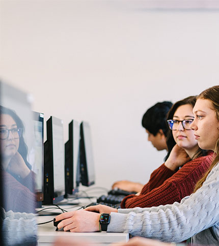 A group of people working at computers in a classroom setting. Two women in focus are looking at their screens, wearing sweaters and glasses. A person in the background is also using a computer. The atmosphere seems studious and focused.
