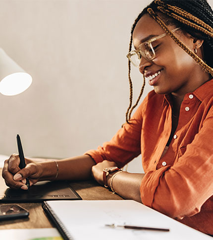 A person with braided hair and glasses is smiling while using a digital pen on a tablet. They are sitting at a desk with a lamp, notebook, and smartphone.