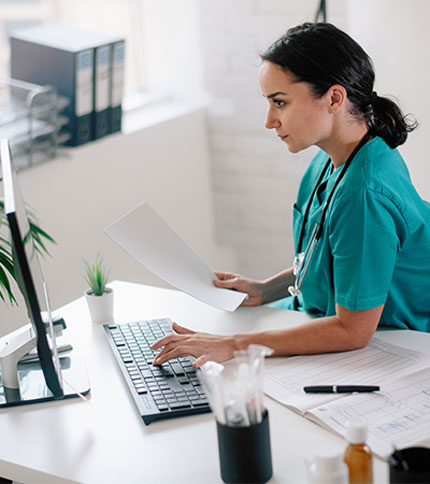 A healthcare professional in teal scrubs sits at a desk, typing on a keyboard while holding a document. A stethoscope hangs around their neck. The desk has office supplies, papers, and a small potted plant. Shelves with binders are seen in the background.