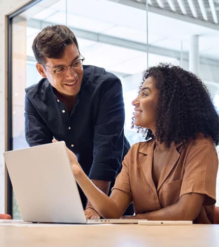 Two colleagues smiling and talking while working together at a desk with a laptop in a modern office setting, demonstrating strong organizational management skills.