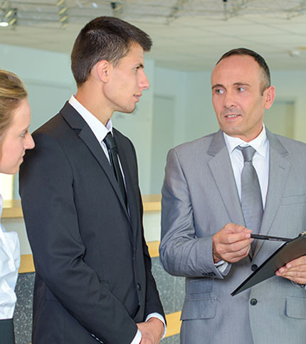 Three professionals in business attire are engaged in a serious discussion. Two men, wearing suits and ties, stand facing each other, while a woman in a white blouse listens attentively on the left. One man is holding a clipboard.