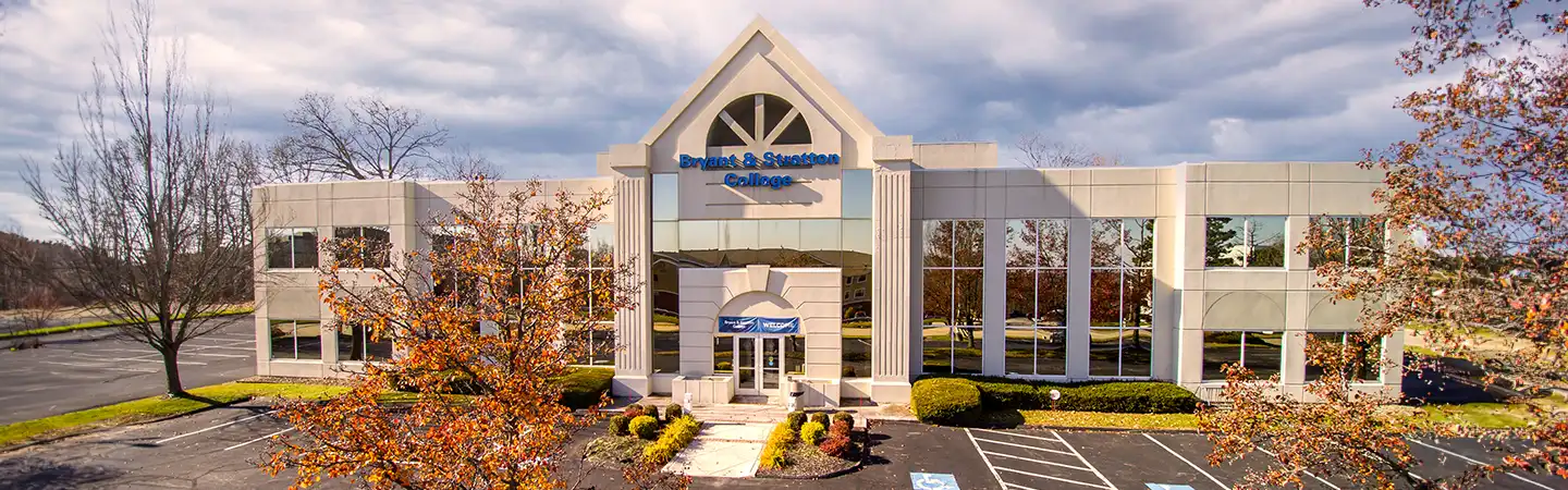 Modern two-story office building with "Dean & Spanton College" sign, large windows, and parking lot surrounded by trees.