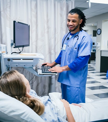 A smiling healthcare professional in blue scrubs stands at a computer workstation, talking to a patient in a bright, clean hospital room—highlighting the caring atmosphere offered by top nursing programs in Rochester NY.