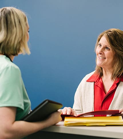Two women are engaged in a conversation at a desk. One has a tablet, and the other has a folder. The background is a plain blue wall, and both appear to be smiling.