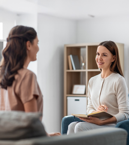 Two women are sitting indoors, facing each other. One woman holds an open notebook and smiles while listening, suggesting a friendly conversation or a counseling session—an example of roles you can pursue with a bachelor’s in social services.