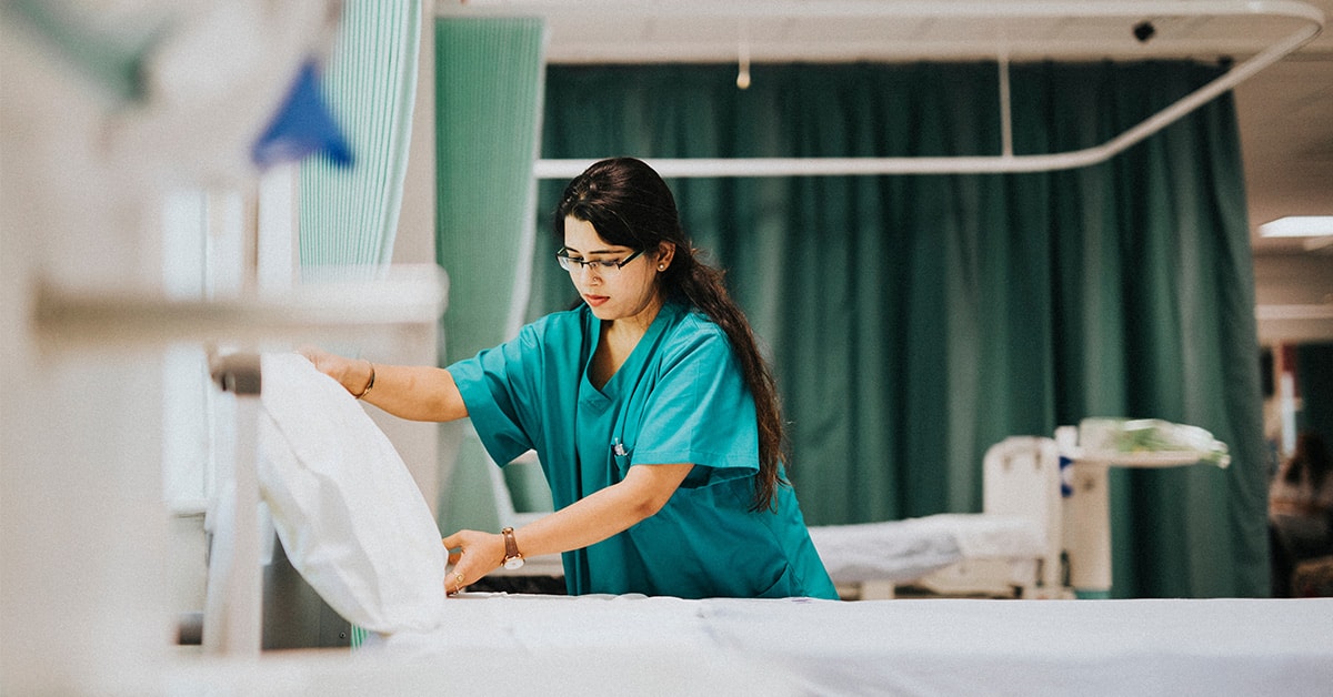 A healthcare worker in green scrubs, likely a BS Nursing generalist, adjusts a pillow on a hospital bed in a room with green curtains. The clean and organized setting suggests a hospital or medical facility environment in OH or WI.