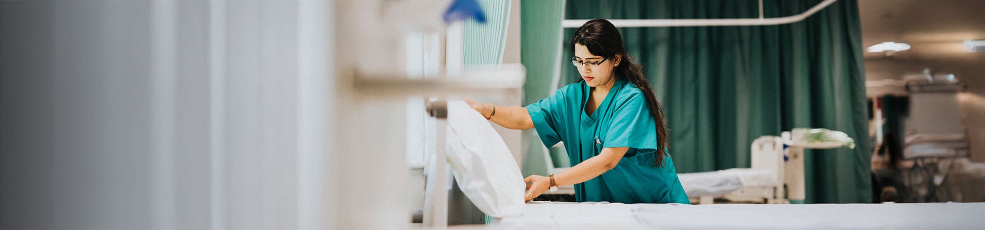 A BS Nursing generalist in teal scrubs neatly makes a hospital bed in a clean, well-lit room with green curtains and medical equipment, showcasing skills valued in OH WI healthcare settings.