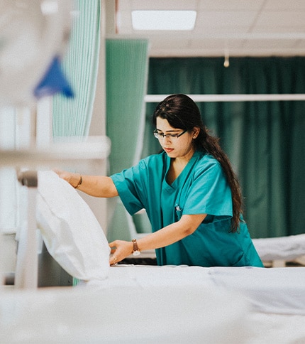 A generalist healthcare worker in teal scrubs, possibly with a BS Nursing from OH or WI, makes a bed in a hospital room, adjusting a pillow. Green privacy curtains and medical equipment are visible in the background.