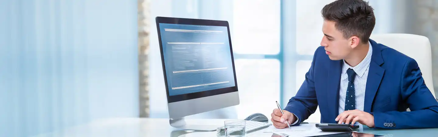Businessman in a blue suit working at a desk with a computer and paper documents, pen in hand, in an office setting.