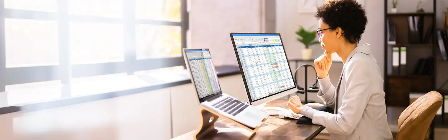 A person works on spreadsheets using a dual monitor setup at a desk, with sunlight streaming in through the window.