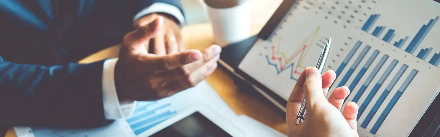 Two people discussing data charts during a business meeting, with graphs and a cup of coffee visible on the table.