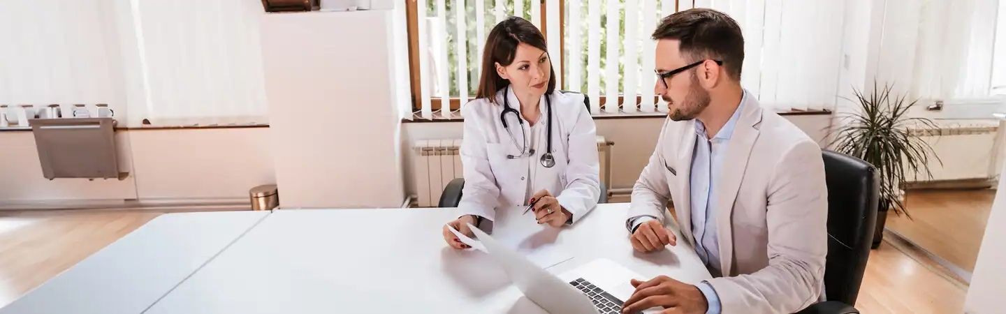A doctor and a man in a suit sit at a white desk, discussing something using a laptop in a bright, modern office.