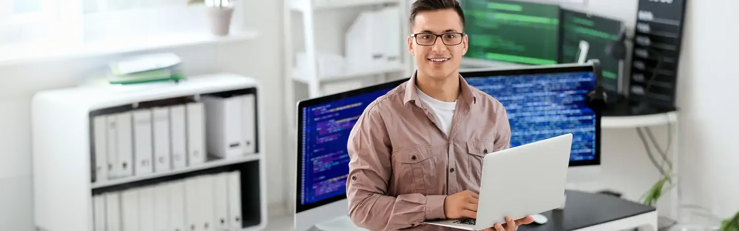 A person wearing glasses and holding a laptop sits in front of monitors displaying code, smiling at the camera.