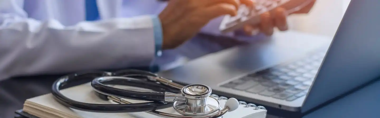 A stethoscope on a notebook beside a laptop, with a person typing on the keyboard in the background.