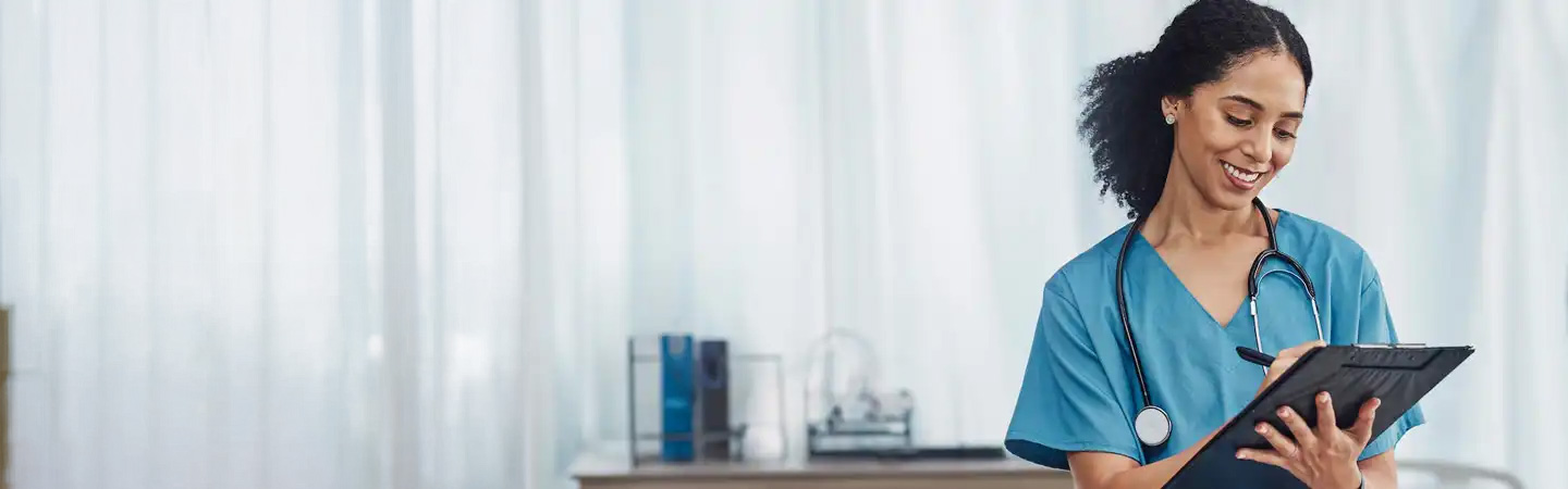 A smiling medical professional in blue scrubs writes on a clipboard with a stethoscope around their neck in a bright room.