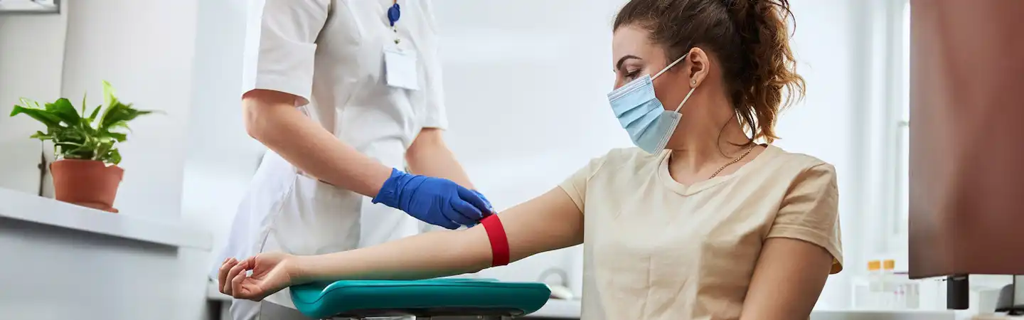 A healthcare worker prepares to draw blood from a masked woman sitting in a medical clinic.
