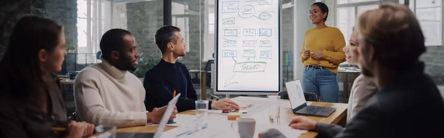 A woman presents a flowchart to a group of colleagues seated around a table in a modern meeting room.